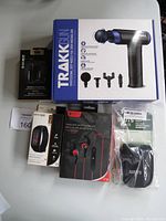 Photo showing boxed Trakk massage gun, Escape wireless earphones, ICEZONE sport earphones, fitness tracker watch, and Roots camera case pouch arranged on table