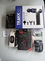 Full view of bundled items including Trakk massage gun box, Skullcandy earbuds, Escape fitness tracker, wireless sport earbuds, and Roots camera case pouch.