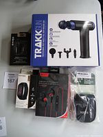 Photo showing the boxed Trakk massage gun, Escape TWS wireless earphones, Escape fitness tracker, red wireless sport earbuds, and Roots camera pouch in original packaging.