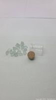 Photo of 15-16 small natural aquamarine crystals, soft blue color, translucent, displayed on white background next to a small clear vial with cork stopper.
