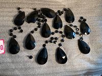 Lot of black glass chandelier prisms in teardrop shape approximately 3 inches long with attached smaller connector beads, displayed on gray surface.