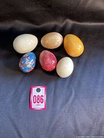 Six decorative eggs on display, including white, beige, orange, cream marble eggs, one red marble egg, and one blue cloisonné metal egg with floral design.