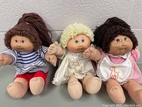 Three 1980s Cabbage Patch Kids dolls sitting together with visible clothing and hair styles showing distinct rag doll and vinyl head features.