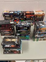 Multiple stacks of assorted movie VHS tapes arranged on a surface showing over 80 titles including X-Men, Jurassic Park, Erin Brockovich, Gladiator, The Italian Job, and others.