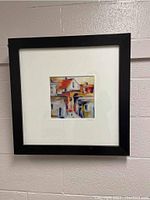 Framed artwork depicting abstract urban buildings with a black frame hung on a cinderblock wall.