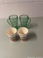 Photo showing two green Coca-Cola glass mugs with handles and two striped paper cups with Canadian Olympic Team Coca-Cola branding on a gray surface.