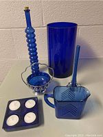 Photo of six cobalt blue glass items arranged on a white surface against a white brick wall. Items include ribbed bottle, tall vase, pitcher, tealight holder, round dish with metal handle, tall slender vase.