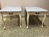 Pair of white end tables with scalloped edges, curved legs, a single drawer with decorative metal handle, placed on terrazzo floor against light tan wall.