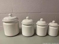 Four white glazed ceramic canisters by Four's Stoneware arranged from largest to smallest with lids on.