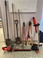 Full set of garden tools leaning against a wall: two shovels, a rake, two pairs of loppers, an axe, electric trimmer and chainsaw visible