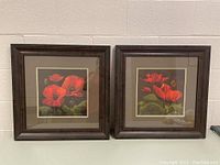 Two framed floral artworks by Gloria Eriksen side by side against a white wall, both with dark wood frames, beige and gold double matting, and bright red poppy flowers.
