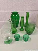 Full view of all eight glass pieces on a white surface against a white wall showing seven green and one clear glass item