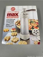 Box showing Cookie Max cookie press, cushion-grip handle, ergonomic design, and 12 unique design discs with images of cookies.