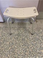 Front view of shower chair showing white plastic curved seat with drainage holes, hand grips, and aluminum legs
