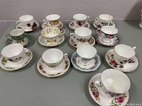 Group photo of 13 assorted vintage teacups with matching saucers featuring various floral and gold designs on a table.