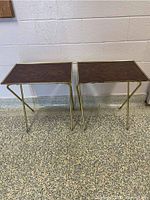 Two vintage folding TV trays standing side-by-side showing gold metal frames and dark brown tabletops with minimal wear.