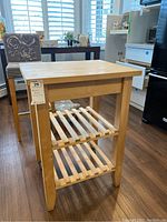 Front view of wooden butcher block table standing on wood floor, with two slatted shelves and caster wheels visible on back legs.