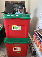 Two red plastic holiday storage totes with green lids, one topped with a box for a Noma swirling snowflake projector.