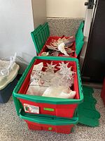Three red and green plastic totes stacked, partially open, showing some white and silver star decorations and a fabric angel figure inside.