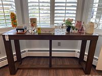 Full front view of the wood console table positioned against a wall with white window shutters. Various decorative items placed on the tabletop, not included in sale.