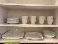 Shelf with four stacked white Corelle side plates, four Corelle mugs with floral design, four Corelle dinner plates with floral design, and four Corelle bowls with green rim stacked.