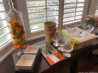 Photo of a clear crystal bowl in box, glass jar with lid containing artificial fruit, ceramic container, glass candle holders, square ceramic dishes, and a decorative notebook on a table near window.