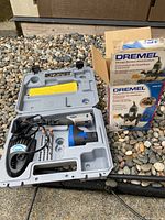 Open grey plastic carrying case containing a cordless Dremel rotary tool, battery charger, battery, and several bits and sanding attachments. Next to the case is a boxed Dremel plunge router attachment.
