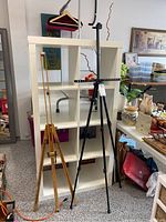 Two adjustable easels shown side by side in front of a white shelving unit; wooden easel on left, black metal easel on right.