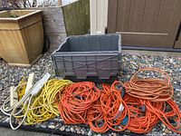Multiple coiled orange extension cords, yellow braided rope, and white power bars next to a plastic bin on ground with rocks.