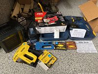 Overview of all items including Skil jigsaw box, Mastercraft drill with battery, Stanley stapler with staples, space heater, telephone, and extra staples on garage floor.