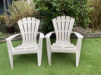 Pair of white resin outdoor Adirondack-style chairs sitting on artificial grass with greenery in the background. Shows general condition and design of both chairs.