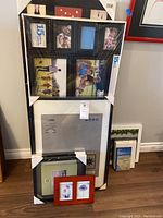 Photo showing a black multifoto frame with six 15x10 cm photo openings, two IKEA picture frames 50x70 cm in original packaging stacked behind, and additional frames including a red double floral frame.