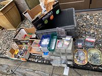 Wide shot of various boxes and plastic organizers with hardware and tools stacked on gravel surface next to a building