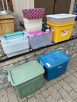 Stack of assorted storage totes and baskets, mostly plastic with lids, some damaged, arranged on stone retaining wall and pavement outside.
