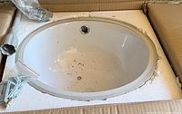 Photo of a new, oval white ceramic bathroom sink in a cardboard box with foam padding, showing included chrome faucet and mounting brackets.