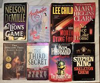 Photo showing front covers of eight used paperback books including titles by Nelson DeMille, Lee Child, Mary Higgins Clark, Stephen King, and others.