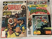 Photo of two comic books: Marvel's Greatest Comics #73 and Adventure Comics #431 featuring Aquaman, showing front covers with wear and creases visible.
