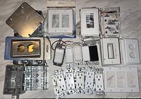 Top view of assorted used electrical switches, cover plates, metal and plastic electrical boxes arranged on a flat surface.