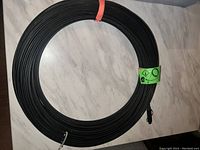 Photo of a black fibre optic cable coil approximately 100 feet in length, coiled neatly on a surface, featuring an orange tie and a green safety label.