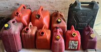 Photo displaying 11 used plastic gas cans of various sizes, mostly red and orange with one dark green. Caps vary in type and color. Some cans show visible dirt and wear.