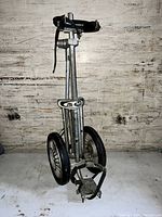 Golf club caddy cart standing upright showing whole frame, two large wheels, adjustable strap, and locking foot pedal.
