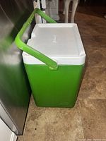 Side angled view of green and white Coleman cooler with handle up, placed on floor.