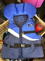 Photo shows a blue youth life jacket by Fluid with adjustable black buckled straps and white reflective stripes, hanging or placed over a crate in a cluttered area.