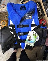 Blue adult large life jacket with three adjustable front buckles, straps, and product tag visible, placed on a surface with other items around.