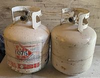 Two used 20lb propane tanks side by side, one with hazard label and both showing paint wear and rust.