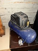 Small blue air compressor with an attached black motor housing sitting on a dark surface, showing surface dust and wear.