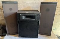 Lot of three used speakers including two floor-standing Polk Audio Monitor 40 speakers and one JBL subwoofer, placed on a wooden surface against a wall.