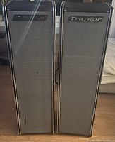 Front view of two tall rectangular floor standing speakers with fabric grilles, slight damage on one grille visible, in black and silver trims.