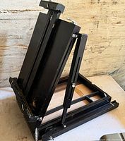 Photo of black wooden easel folded and standing on a flat surface, showcasing its foldable legs and adjustable parts.