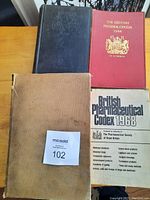 Top view of three manuals on wood table: dark blue, red British Pharmacopoeia 1948, and beige with wear
